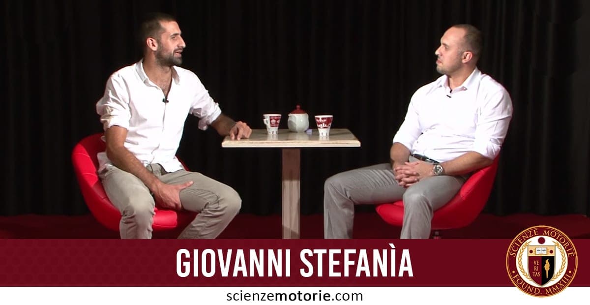 Giovanni Stefanià e Giacomo Catalani seduti su sedie rosse di fronte a un tavolino con tazze e una teiera, entrambi in camicia bianca e pantaloni chiari, con tenda scura sullo sfondo.