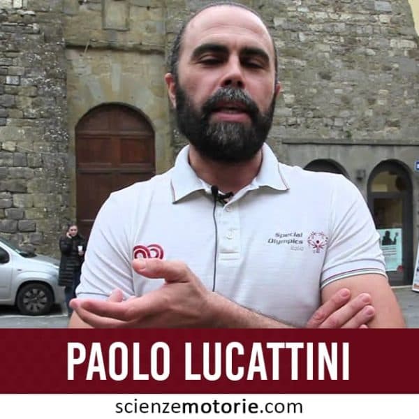 Uomo con barba in polo bianca davanti a un edificio di pietra con persone e una macchina parcheggiata, Paolo Lucattini.