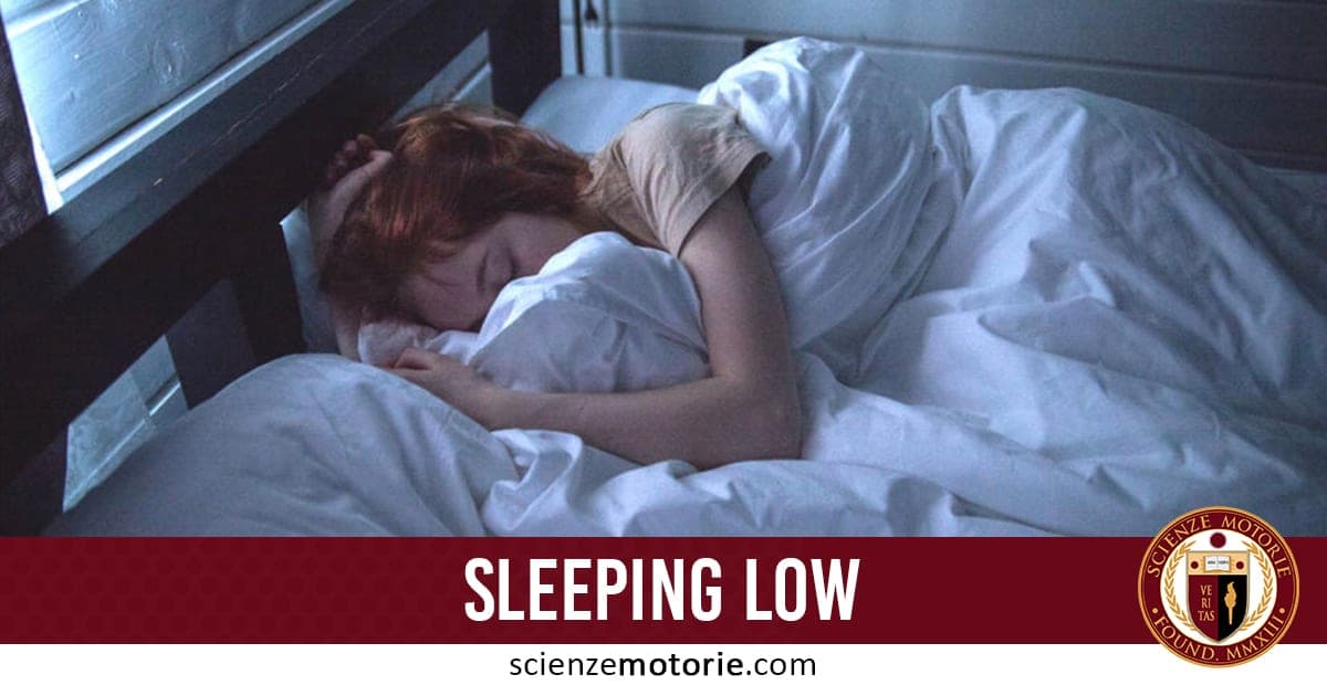 Persona con capelli rossi dorme sotto una coperta bianca in una stanza poco illuminata, con scritta “SLEEPING LOW” e logo di scienzemotorie.com.